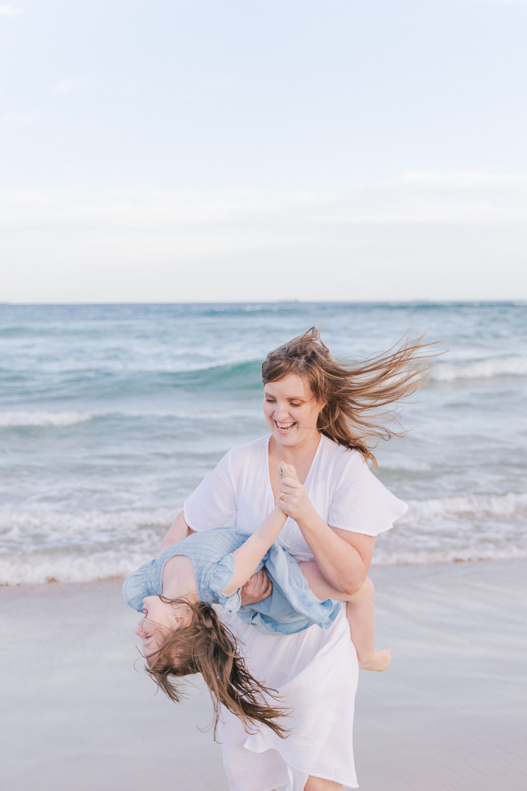 Mum wearing a white dress laughing and spinning her daughter around on the beach