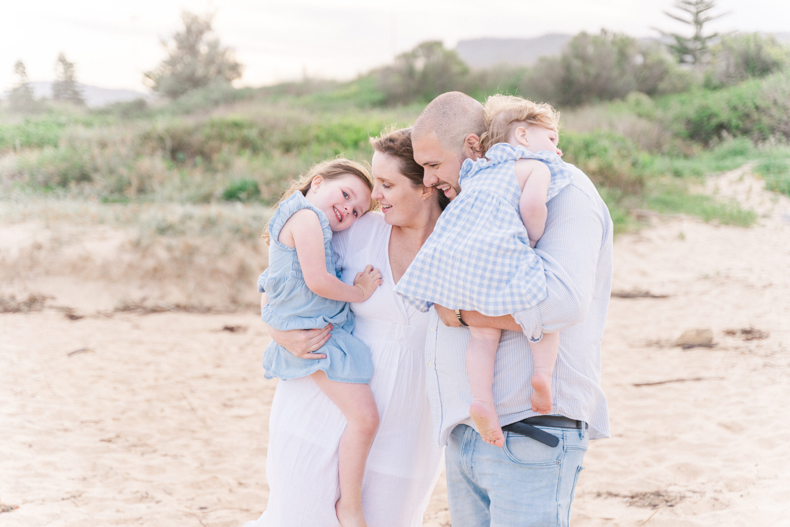 Family of four dressed in blue and white coordinating outfits snuggling on the beach during their family photo session with Stacey McCarthy Photography
